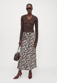 Rjava čipkasta bluza z dolgimi rokavi in scalloped robovi, kombinirana z dolgim krilom z belim in rjavim vzorcem. Dodatki vključujejo sončna očala in majhno torbico.