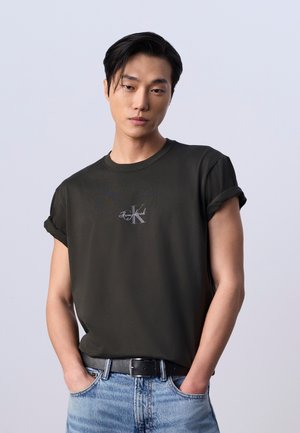 Jeune homme aux cheveux foncés portant un t-shirt noir aux manches retroussées et un jean bleu, debout les mains dans les poches devant un fond uni.
