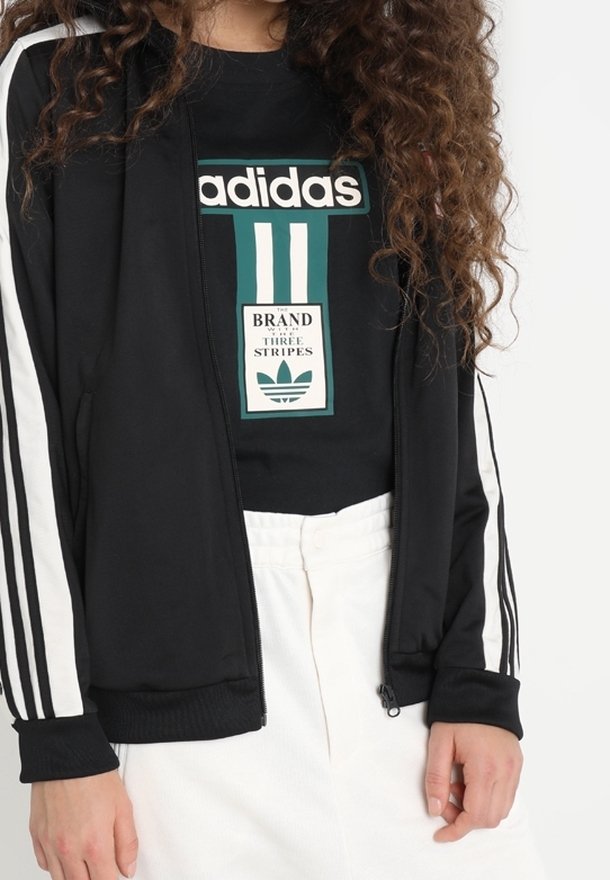 Persona con una chaqueta negra de Adidas con rayas blancas en las mangas y una camiseta negra de Adidas con el logo y texto en verde y blanco.