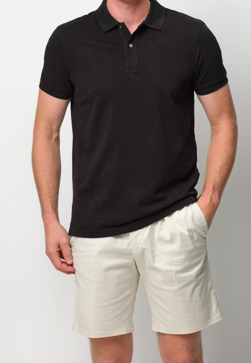 Homme portant un polo noir à manches courtes et un short beige clair, une main dans la poche, debout devant un fond uni.