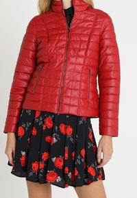 Rød quilted puffer jakke med høj krave, frontlynlås og to lynlåslommer på siden, båret over et sort blomster mønstret nederdel.