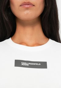 Balta medvilninė marškinėlių su apvalia iškirpte, su juodu stačiakampiu grafišku piešiniu, kuriame rašoma "KARL LAGERFELD JEANS" ryškiu šriftu.