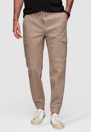 Homme portant un pantalon jogger cargo beige avec des poignets élastiques, des baskets blanches, une chemise noire et des bracelets noirs, debout les mains dans les poches.