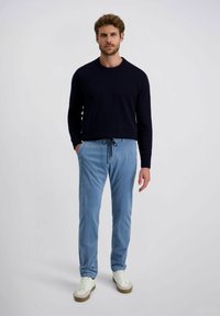 Man met krullend haar in een zwarte trui, lichte blauwe sweatpants met touwtjes en witte sneakers, staande tegen een effen achtergrond.