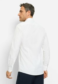 Weißes Langarmhemd mit strukturiertem Muster, Knopfmanschetten und spitzem Kragen, getragen mit marineblauen Hosen, von hinten betrachtet.