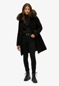 Svart puffjacka med en huva fodrad med päls, dragkedjestängning och sidofickor. Bärs över en mörk stickad tröja och slim-fit svarta jeans med snörkängor.