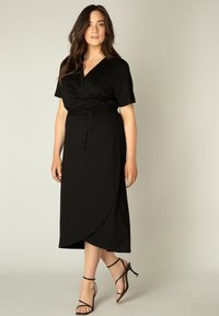 Zwarte wikkeljurk met korte mouwen en een strikceintuur op de taille. Semi-getailleerd ontwerp, knielang, van zachte stof, gedragen met zwarte sandaaltjes met hakken.