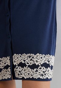 Marinblå klänning med en slät textur, prydd med en vit blommig spetskant och knappar längs framsidan. Mjukt tyg och knälång design.