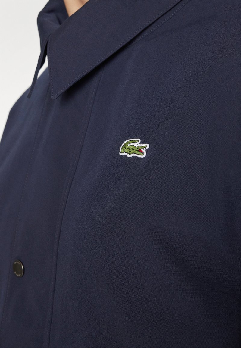 Camicia blu navy con colletto a punta e un piccolo logo di coccodrillo verde ricamato sul petto vicino ai bottoni neri.