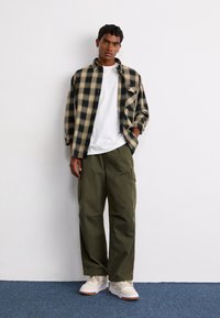 Plaid oversized shirt in black and beige, white t-shirt underneath, olive cargo pants, and cream sneakers with gum sole, standing against a wall.