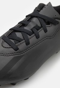 Botas de fútbol negras con parte superior sintética lisa, cordones texturizados, una marca sutil y una suela moldeada con tacos cónicos.