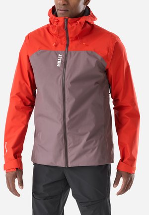 Homme portant une veste à capuche zippée rouge et grise avec un pantalon noir, debout devant un fond blanc uni.