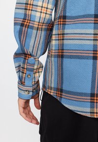 Blauw geruit shirt met oranje en zwarte checkpatronen. Gestructureerde stof met knoopaccenten bij de manchetten. Zoom is gebogen.