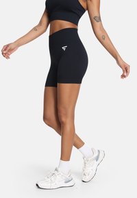 Crne visoke biciklističke kratke hlače s glatkom teksturom, s diskretnim logotipom. Uparene s bijelim tenisicama i čarapama do članaka.