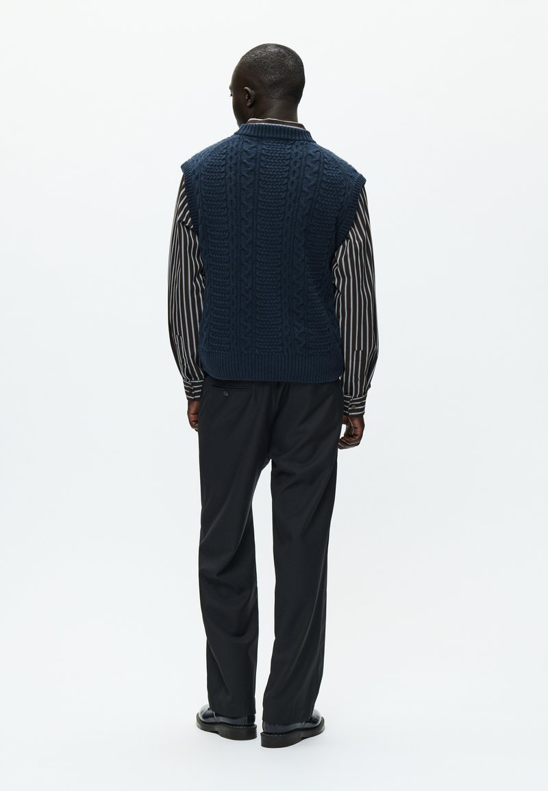 Gilet sans manches en tricot bleu marine avec motif en cable, porté par-dessus une chemise rayée, associé à un pantalon noir. Texture douce, coupe décontractée.