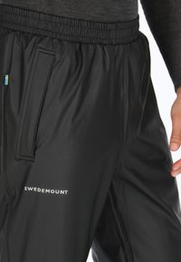 Zwarte waterdichte broek met een elastische tailleband, voorzien van een zijzak en het logo "SWEDEMOUNT". Inclusief een klein accent van de Zweedse vlag.