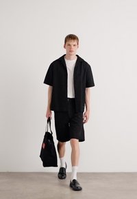 Cămașă neagră cu mânecă scurtă și guler, cu un model texturat, asortată cu pantaloni scurți negri. Modelul ține o geantă de umăr neagră, un tricou alb și șosete.