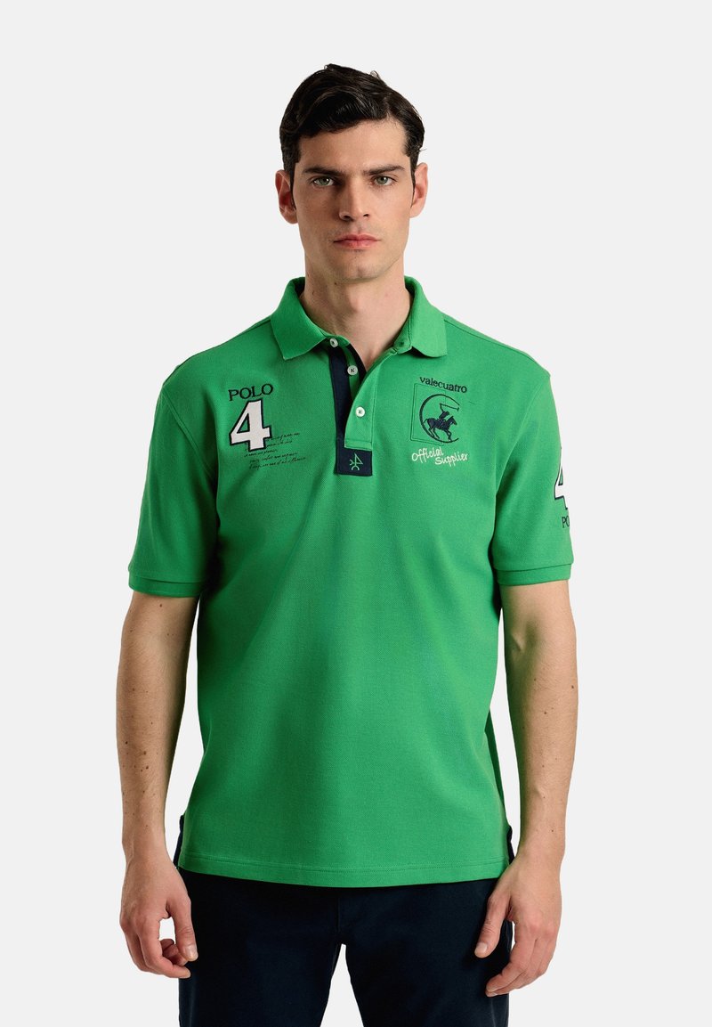 Polo verde con cuello azul marino, que presenta un logotipo, el número "4" y texto bordado en el lado izquierdo y la manga. Hecho de tejido texturizado.