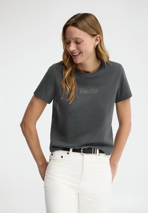 Young woman smiling, wearing a dark gray "Polo Club" t-shirt, white high-waisted jeans, and a black belt with gold buckle.