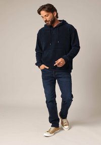 Sweatshirt à capuche bleu marine avec poche kangourou, associé à un jean bleu foncé et des baskets beige. Texture douce et coupe décontractée.