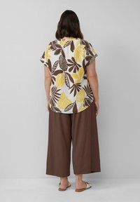 Bluse mit floralem Muster, kurzen Ärmeln und gelben sowie braunen Blättern, kombiniert mit weiten braunen Hosen und schlichten Sandalen. Rückansicht.