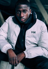 Lichtgrijze pufferjack met elastische manchetten, gedragen over een zwart sweatvest met rits. Prominent logo op de linkerkant van de borst van de jas. Donkere achtergrond.