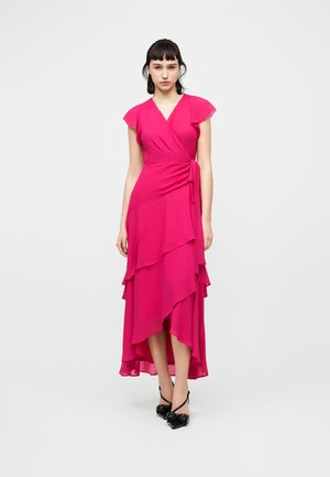 Kobieta stoi w sukience kopertowej w kolorze fuksji z falbankowymi rękawami i asymetrycznym, warstwowym dołem, nosząc czarne szpilki z zaostrzonym czubkiem.