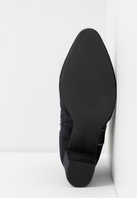 Enkel zwart suede puntige schoen, schuin met de textuur van de zool naar voren gericht tegen een witte hoek achtergrond.