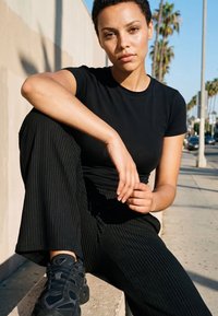 Fiatal nő rövid, göndör hajjal, fekete ingben és nadrágban, egy kültéri betonperemen ülve, háttérben pálmafák és utca látható.