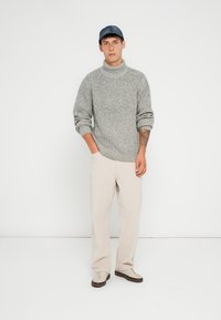 Maglione grigio con texture e colletto alto a coste, abbinato a pantaloni beige chiaro e sneakers chiare, completato da un cappellino blu.