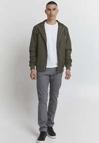 Veste légère à capuche vert olive associée à un T-shirt blanc, un pantalon slim gris et des baskets noires. Design simple, sans motifs.