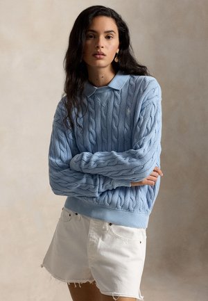 Donna con lunghi capelli scuri che indossa un maglione blu a trecce sopra una camicia con collo e pantaloncini di denim bianchi su sfondo neutro.