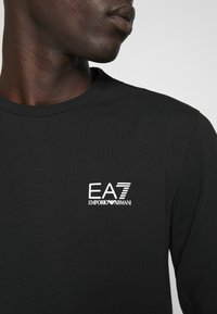 Maglietta nera a maniche lunghe realizzata in tessuto morbido, con il logo bianco EA7 Emporio Armani sulla parte sinistra del petto.
