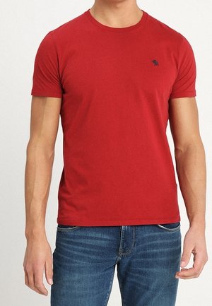 Mann trägt ein schlichtes rotes T-Shirt mit kurzen Ärmeln und blaue Jeans, steht vor einem schlichten hellen Hintergrund.