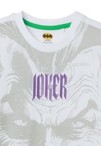 Camiseta blanca con cuello verde que presenta un gráfico del Joker en púrpura y gris. El diseño incluye intrincados detalles faciales y trabajo de líneas.