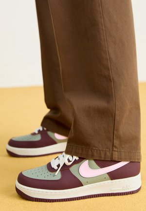 Person iført brune bukser og flerfarvede sneakers med pink Nike-swoosh, hvide snørebånd og beige såler, der står på gul gulv.