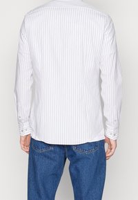 Bílá košile s dlouhým rukávem a tenkými vertikálními pruhy, s rovnými lemy a knoflíkovými manžetami, kombinovaná s modrými džínovými kalhotami.