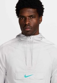 Nike lichtgewicht hoodie in lichtgrijs. Voorzien van een buidelzak met turquoise logo, ritsluiting en textuurstof.
