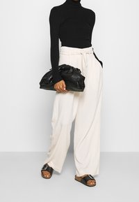 Col roulé noir côtelé, pantalon large crème, et sac à main noir texturé. Sandales avec boucle argentée et semelle en liège. Fond neutre.