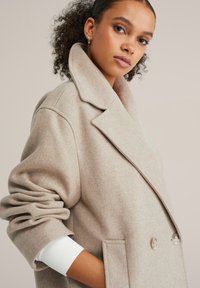 Ženska s kodrastimi lasmi, oblečena v bež prevelik plašč z širokimi ovratniki, z rokami v žepih in zlatimi obročastimi uhani na nevtralnem ozadju.