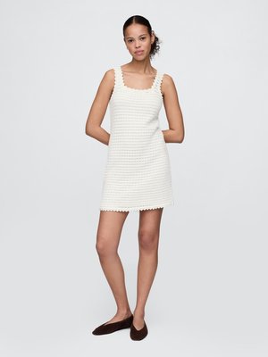 Woman standing with arms behind back wearing sleeveless textured white dress and brown flat shoes against plain background.