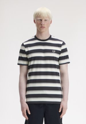 Ung mand med lyst blondt hår iført en sort og hvid kortærmet skjorte med vandrette striber og mørke bukser på en ensfarvet baggrund.