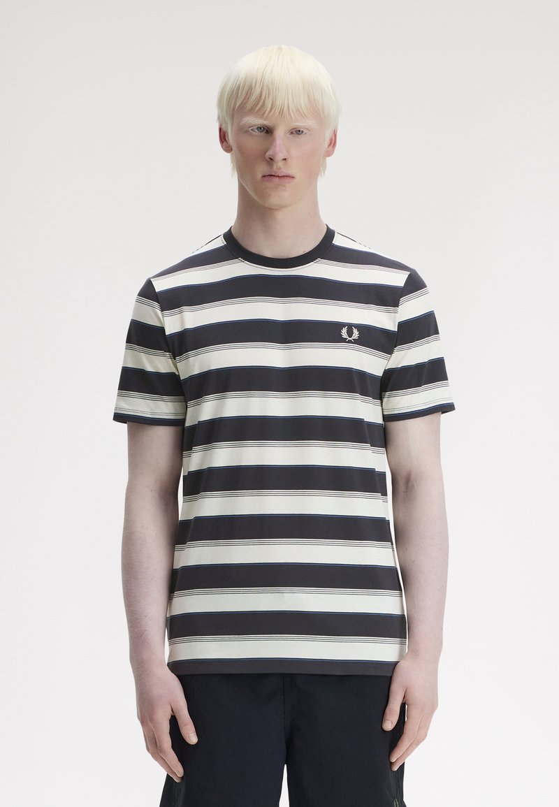 Joven con cabello rubio claro, que lleva una camiseta de manga corta con rayas horizontales en blanco y negro y pantalones oscuros sobre un fondo liso.