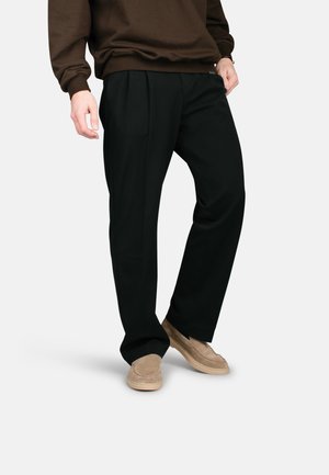 Persoon met zwarte geplooide broek, bruine trui en beige suède instapschoenen, staand met één hand in de zak tegen een witte achtergrond.