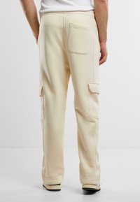 Pantalone od dukserice boje krema s opuštenim krojem, elastičnim pojasom, bočnim cargo džepovima i vidljivim detaljima šavova. Mekana, teksturirana tkanina.