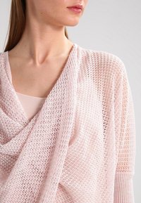 Hellrosa offene Strickjacke mit hervorgehobenem Vorderteil, gekämmten Bündchen und strukturiertem Muster, getragen über einem passenden Oberteil.