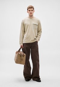 Béžová košeľa s dlhými rukávmi s náprsným vreckom, v kombinácii s hnedými nohavicami s širokými nohavicami, držiaca béžovú tašku s hnedými akcentmi, na pozadí sivej steny.