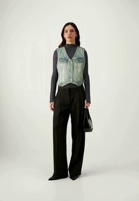 Colete de ganga com lavagem clara, apresentando bolsos frontais e botões metálicos, sobre uma blusa preta de manga longa. Combinado com calças pretas de perna larga.