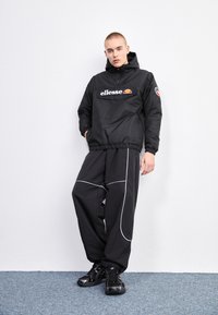 Giacca a vento nera con cappuccio e logo sul davanti, abbinata a pantaloni neri larghi con piping bianco e sneakers nere.
