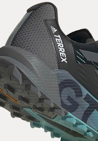 Scarpa sportiva nera con una parte superiore in rete testurizzata, caratterizzata da una suola grigia con battistrada pronunciato e accenti blu. Logo in evidenza.